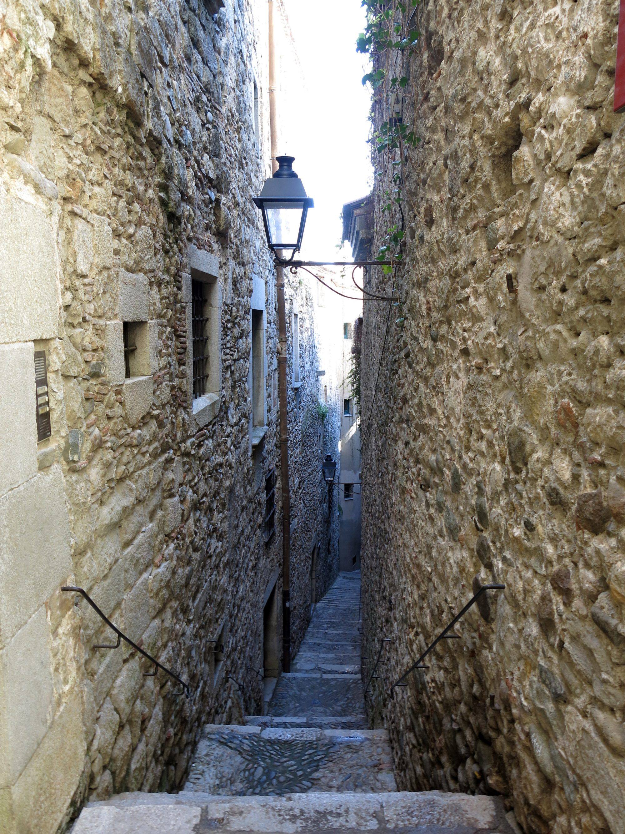 Carrer de Sant Llorenç in the Call, the medieval Jewish quarter of Girona: a very narrow stepped lane between tall rubble-stone walls, with iron handrails on both sides, a wrought-iron street lamp hanging overhead and the steps running down into the shade