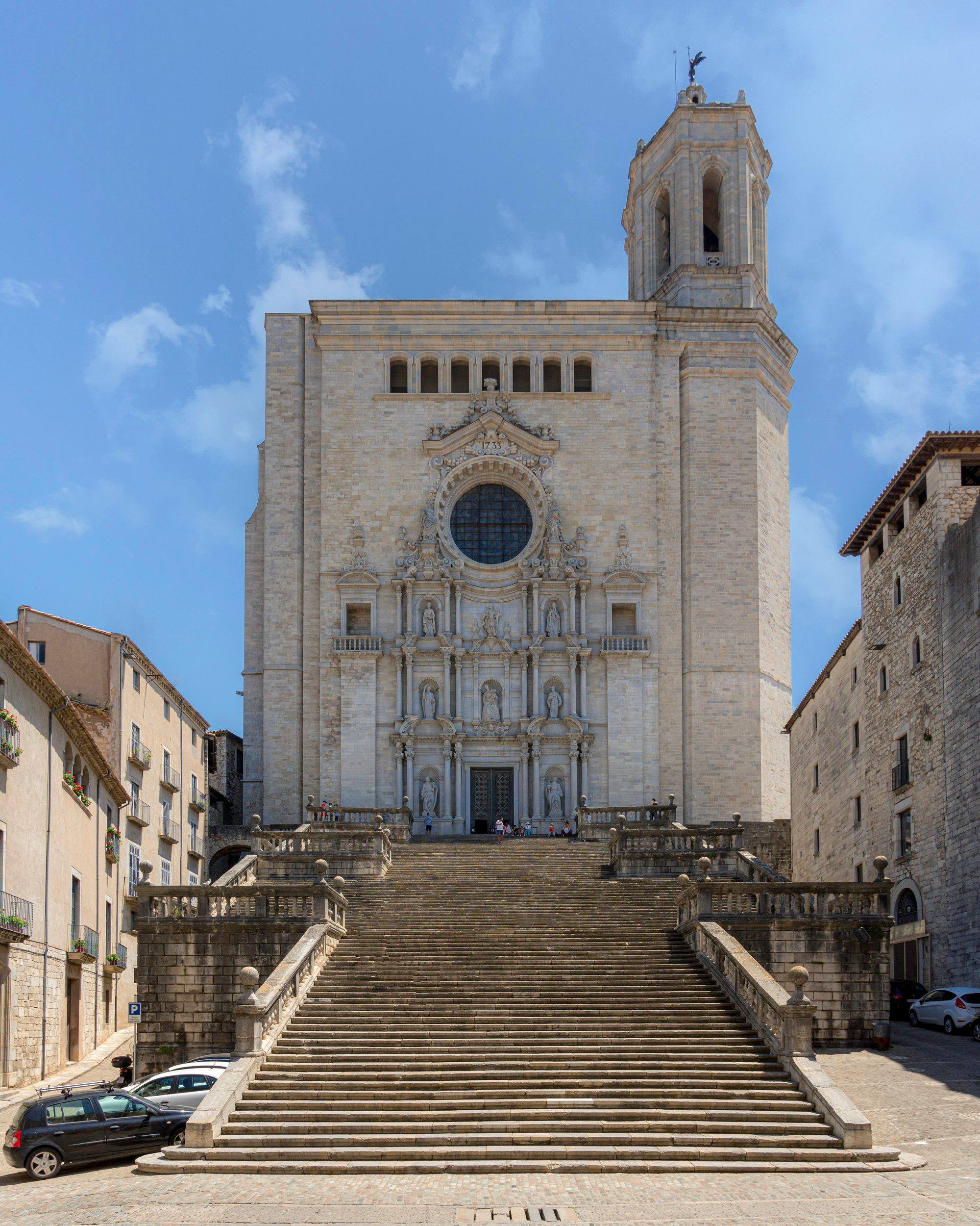 The Cathedral of Girona seen from the Plaça de la Catedral: a broad baroque stairway with stone balustrades climbs in long flights to the baroque main façade with its rose window and statues, the bell tower rising on the right, framed by old stone buildings under a blue sky with a few clouds