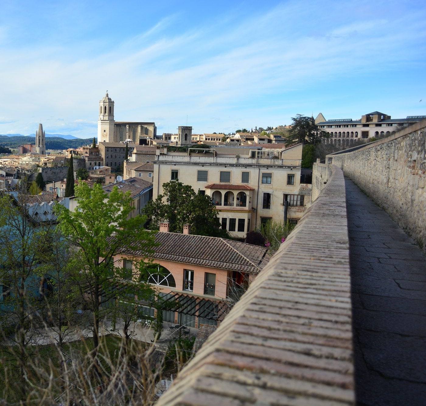 The walkway along the top of Girona's medieval city walls near Casa Crous: the brick-capped parapet runs off on the right while the old town spreads out below among spring trees, with the bell tower of the Cathedral, the Gothic spire of Sant Feliu and wooded hills in the distance under a pale blue sky