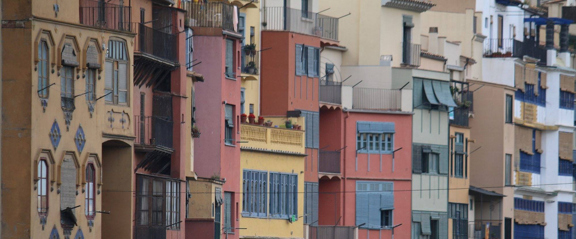 Close-up of the riverside façades of the Cases de l'Onyar: tall narrow houses in ochre, salmon, terracotta, saffron and sage stacked side by side, rows of windows with blue and green roller blinds, iron balconies and an ochre house patterned with blue diamonds