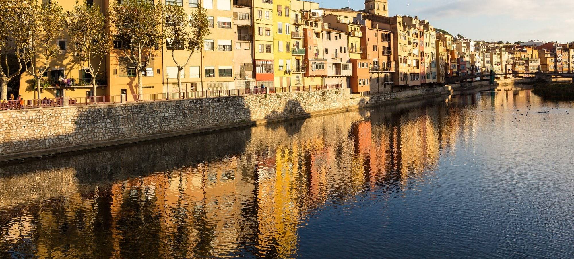 The Cases de l'Onyar in Girona in low golden afternoon light: a row of ochre, saffron, yellow and salmon houses behind autumn trees and a stone embankment, the row receding upstream, their colours mirrored in the calm water of the river Onyar