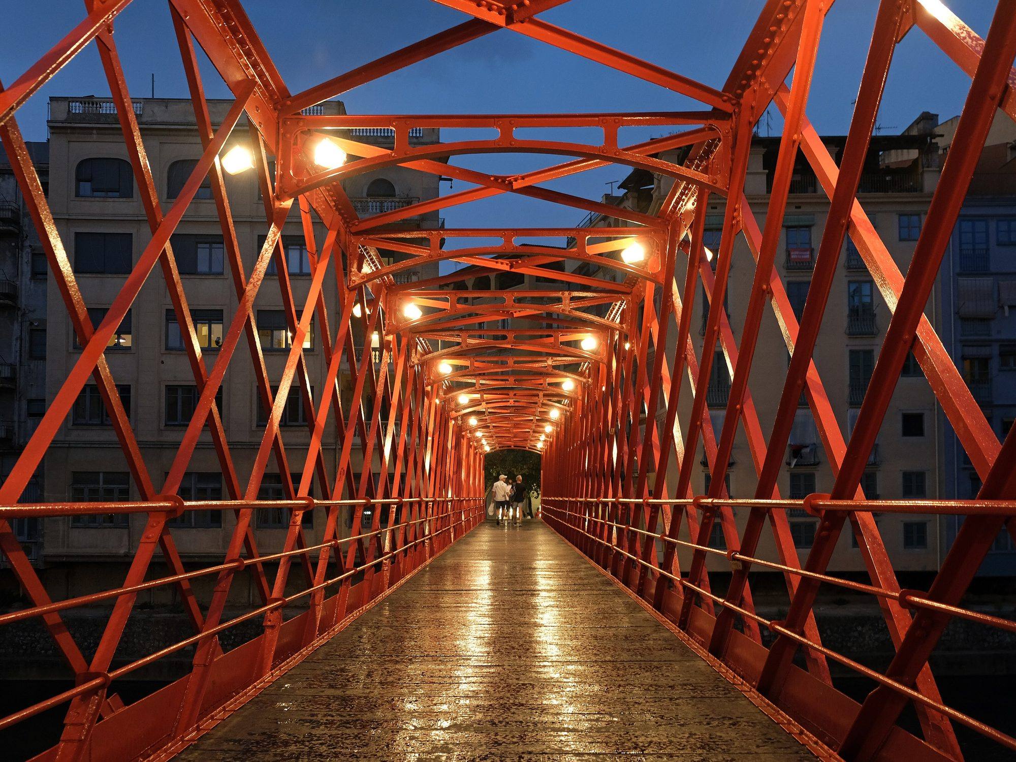 Inside the red iron Pont de les Peixateries Velles, the 1877 Eiffel bridge over the Onyar, at dusk: a tunnel of riveted, criss-crossed red girders lit by lamps, the rain-wet plank deck gleaming as it runs to the far end, and pale riverside buildings behind the lattice against a deep blue sky